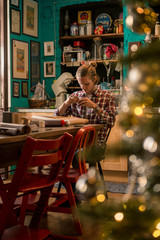 Young adult man with long blonde hair wrapping christmas present with red ribbon seated on wooden table in cozy apartment indoor.