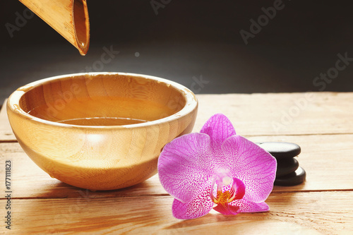 Zdjęcie XXL Miska, która wylewa wodę z łodygi bambusa, kwiatów orchidei i kamieni na gorący masaż na drewnianym stole