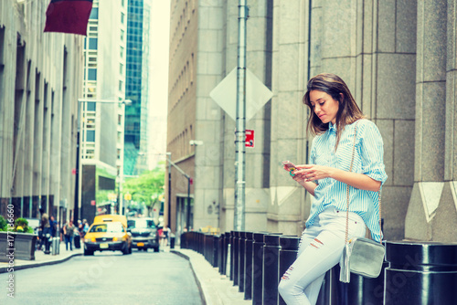 Fotografie, Tablou Young Argentine Woman wearing light blue striped shirt, white destroyed jeans, shoulder carrying small bag, sitting on street in New York, looking down, texting on cell phone