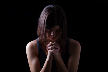 Wall Mural - Faithful athletic woman praying, with hands folded in worship to god, head down and eyes closed in religious fervor, on low key black background. Concept for religion, faith, prayer and spirituality
