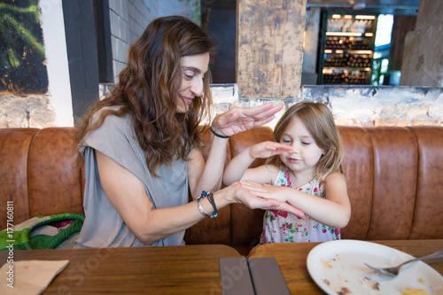 Plakat Cztery lata wiekowa blondynki szczęśliwa dziewczyna bawić się z rękami z kobiety matką i uśmiechniętym obsiadaniem w brown rzemiennej kanapie przy restauracją