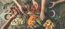 Flat-lay Of Friends Hands Eating And Drinking Together. Top View Of People Having Party, Gathering, Celebrating Together At Wooden Rustic Table Set With Different Wine Snacks And Fingerfoods