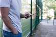 © Maskot - Midsection of young man standing against fence while using smart phone