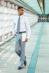 Young African American Businessman working in New York, wearing white T shirt, light gray pants, black leather shoes, tie, holding laptop computer, standing on walkway in office building, looking away