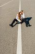 © Iryna - Pretty brunette hipster traveller girl sitting on the middle of asphalt road. Young woman sitting on the high way. Female sitting on center mall of the road. One white line along road.