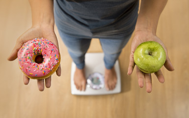  close up woman on weight scale holding in her hand apple fruit and donut as choice of healthy versus unhealthy food