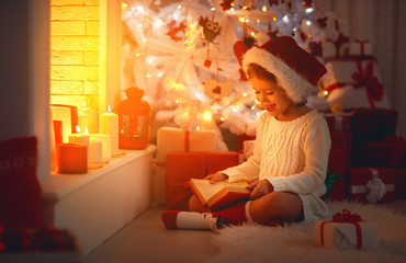 Wall Mural - happy child girl read book at christmas near fireplace at home.