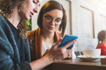 Two young women sit in a cafe at the table and use a smartphone. The girl shows her friend a picture on the phone screen. Social media, network. Girls working together, blogging, checking email.