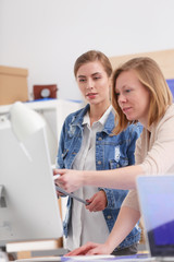 Two young woman standing near desk with instruments, plan and laptop.