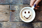Happy face on cappuccino foam, woman hands holding one cappuccino cup on wooden table