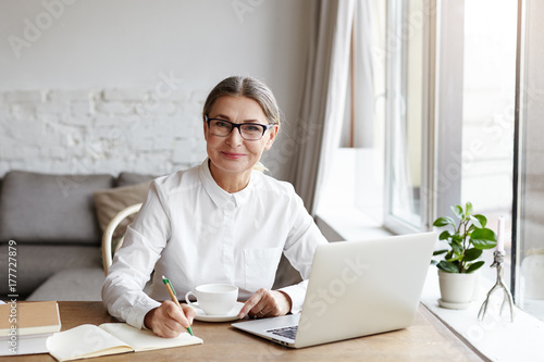 Fotografie, Obraz Attractive positive talented mature woman writer sitting in front of laptop and writing her new book about love, looking at camera with joyfull smile