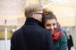 © shurkin_son - Sweet moment of true love. Romantic young couple in winter clothes cuddling outdoors: Hispanic girl with nose ring and blonde Caucasian guy in eyeglasses hugging on street decorated with light garland