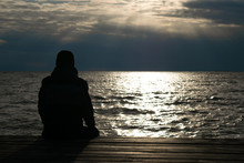 Silhouette Of Lonely Man In Jacket And Cap Who Seating On Wooden Dock And Looking On Sunset Over Sea. Cold Autumn.