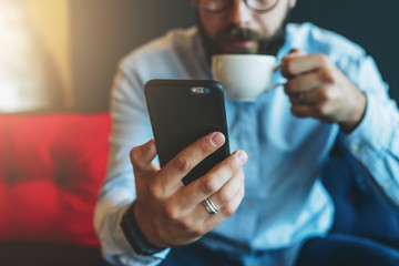 Wall Mural - Close-up of black smartphone in man's hand. In background, in soft focus, young bearded businessman drinks coffee. Guy uses digital gadget, checking email, reading, blogging. Online working,education.