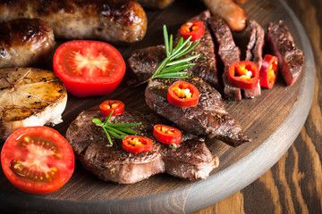 Close-up photo of mixed grilled meat platter. Beef, pork, poultry, sausages, grilled garlic, chili pepper, red tomatoes on wooden rustic background.