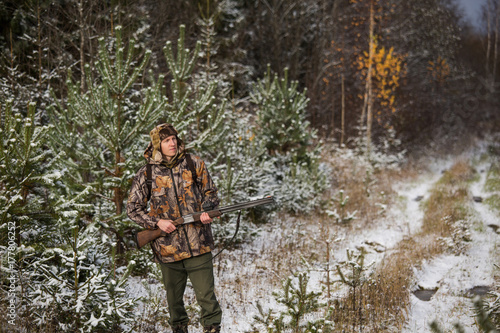 Plakat Mężczyzna myśliwy z plecakiem, uzbrojony w karabin, stojąc w lesie mroźną zimę. Mężczyzna odwraca wzrok. Opadów śniegu w lesie.