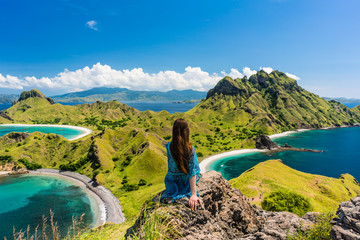 Rear view of a young woman enjoying the awesome view of Padar Island, while sitting on the top of a volcanic mountain during summer vacation in Indonesia