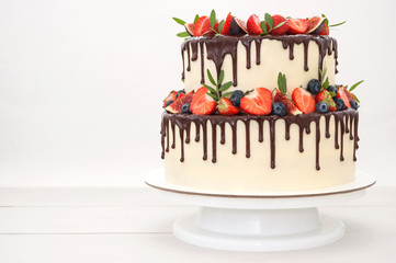 Two-tiered cake in chocolate, decorated with slices strawberries, blueberries, figs and green leaves on a white wooden table. Picture for a menu or a confectionery catalog.