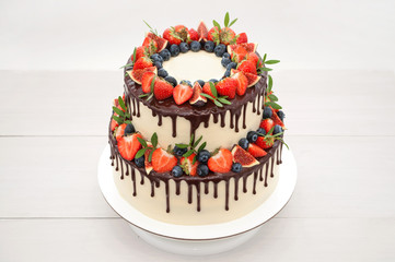 Wall Mural - Two-tiered cake in chocolate, decorated with slices strawberries, blueberries, figs and green leaves on a white wooden table. Picture for a menu or a confectionery catalog.