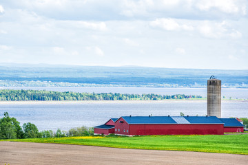 Aerial cityscape landscape view of farmland in Ile D'Orleans, Quebec, Canada, plowed field, furrows, farm and grain storage silo barn, shed or by Saint Lawrence river with hills, mountains and village