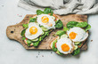 Healthy breakfast sandwiches. Bread toasts with fried eggs and fresh vegetables on wooden board over grey marble background, top view. Clean eating, healthy, weight loss, detox food concept