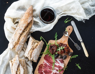 Poster - Wine appetizer set: vintage dinnerware, french baguette broken into pieces, dried tomatoes, olives, smoked meat and arugula on rustic wooden board over dark background. Top view 