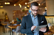 © pressmaster - Waist-up portrait of concentrated bearded manager in eyeglasses checking business emails on digital tablet while standing at spacious modern restaurant