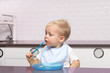 Cute toddler in a blue bib eating banana in the modern kitchen