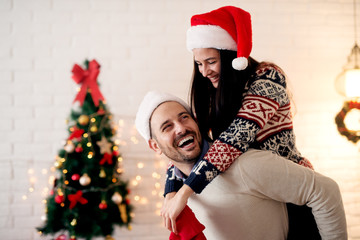 Romantic young couple enjoying Christmas time. Handsome smiling man giving piggyback to his wife at home for Christmas holidays.