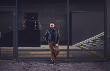 Bearded man in a leather jacket posing near a glass building