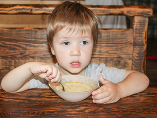 The child sits at the table and eats from a plate on their own. Little boy eats from a plate. The child eats with a spoon..