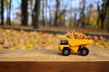 The Concept Of Seasonal Harvesting Of Autumn Fallen Leaves Is Depicted In The Form Of A Toy Yellow Truck Loaded With Leaves Against The Background Of The Autumn Park