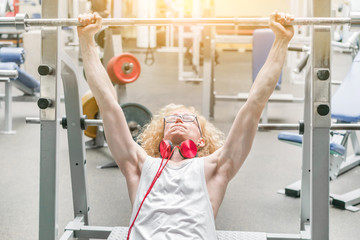 Curly blond man in a white vest with red headphones lifting barbell with effort in the gym