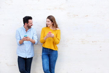 Laughing couple standing by white wall holding mobile phones