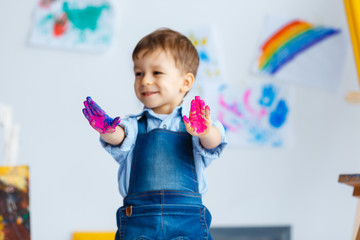 Wall Mural - Cute, happy, white, three year boy in blue shirt and jeans showing his dirty hands in watercolors. Little child having fun after painting. Concept of early childhood education, happy family, parenting