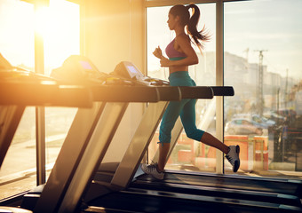 Wall Mural - Young healthy athletic woman running on a treadmill near the sunny window in the gym and listening music.