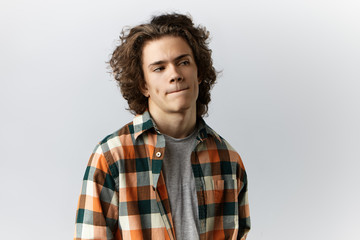 Wall Mural - Headshot of trendy looking young male with dimples and wavy hairstyle looking sideways and pursing lips while thinking over something, feeling uncertain about making important decision or choice