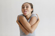 People, human expressions, gesture and body language. Funny young brunette female in grey t-shirt posing at studio wall sticking out her tongue, keeping arms wrapped around herself and eyes popped out