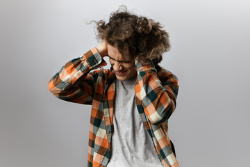 Wall Mural - Picture of annoyed young male wearing stylish checkered shirt over grey t-shirt, grimacing, having painful frustrated look, squeezing head and covering ears, can't stand irritating sound or noise