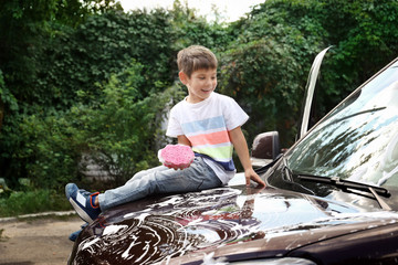 Sticker - Adorable little boy washing car with sponge while sitting on hood outdoors