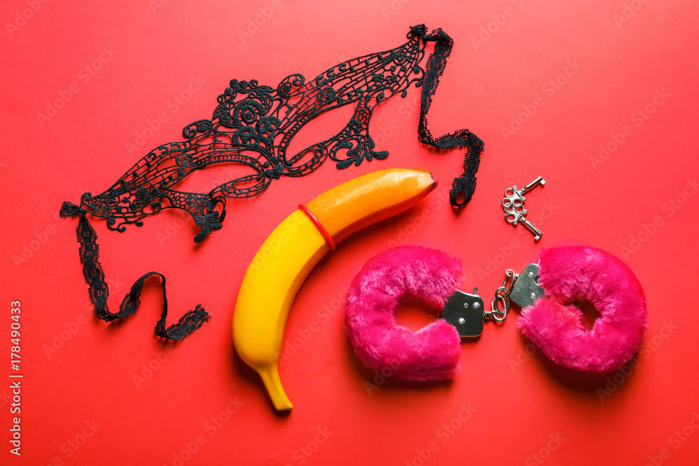 Banana with condom, mask and handcuffs on color background