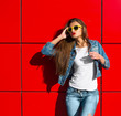 © zadorozhna - Close up fashion lifestyle portrait young hipster girl, wearing bright make up and yellow sunglasses, making sexy faces. Urban red  wall background.