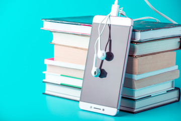 phone with white earphones next to a stack of books on a blue background. concept of audiobooks and 