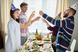 Fototapeta Na drzwi - Group of happy young people wearing holiday caps celebrating Birthday with friends clinking champagne glasses and toasting during dinner party at home