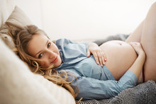 Woman Enjoying Her Pregnancy Relaxing In Comfy Bed On Early Morning. Portrait Of Cheerful Light-haired Future Mother In Cozy Home Clothes, Holding Her Stomach With Hands.