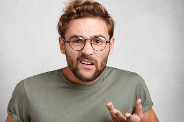 Wall Mural - Portrait of nervous unshaven man gestures and frowns face, looks displeased or dissatisfied, wears spectacles and casual t shirt. Caucasian man with astonished expression poses in white studio