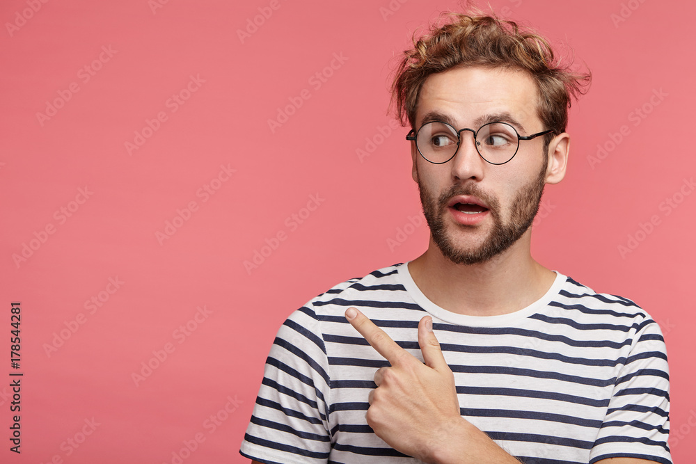 Shocked bearded young man with trendy hairdo and mustache points at copy space as poses against pink background, advertises something or being surprised to see high prices. It`s unbelievable! - obrazy, fototapety, plakaty 
