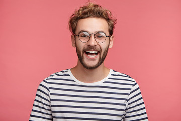 Wall Mural - Cheerful hipster guy smiles happily, has excited expression, dresssed casually, celebrates his anniversary or promotion at work, isolated over pink studio background. People, youth, emotions concept
