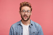 Unhappy handsome bearded young male frowns face in displeasure, looks directly into camera, wears spectacles and denim shirt, expresses hesitation or doubt as being asked disputable question