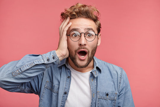 Shocked bearded man with jaw dopped out and bugged eyes, stares at camera as sees awful car accident, being horrified and in stupor. Amazed stressful male adult poses over pink wall indoors.